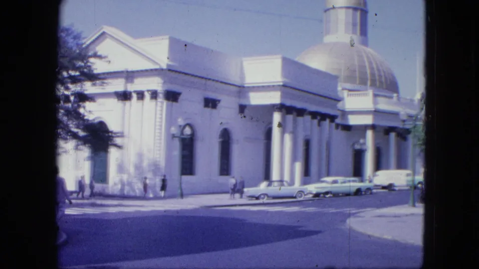 1964 8mm wide view of a large domed public-looking building with columns, arched windows, pedestrians, parked cars, and tree shadow.