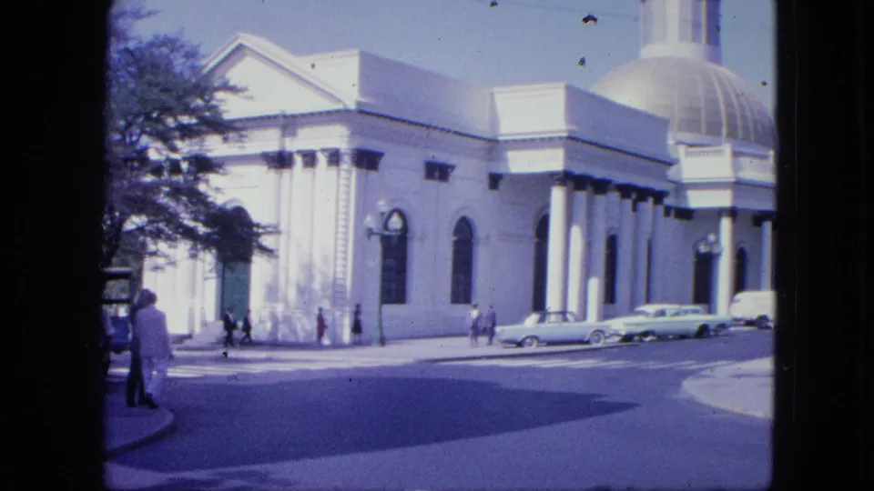 1964 city-street frame showing the domed columned building from a corner angle, with pedestrians, vintage cars, tree shadow, and bright sky.
