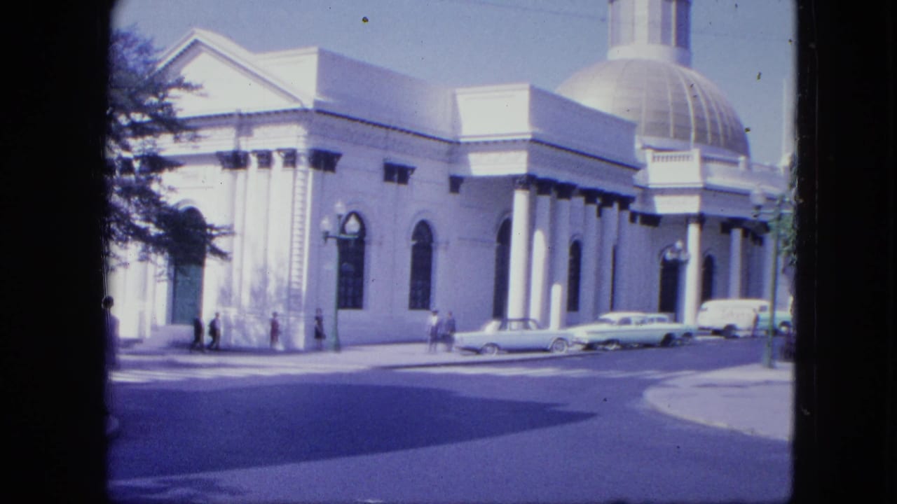 1964 Venezuela street view with a white columned building, dome, arched windows, parked sedans, and a few pedestrians.