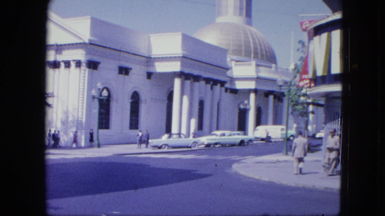 1964 Venezuela street corner showing the white domed building, parked cars, a passing pedestrian, and a striped storefront awning at right.