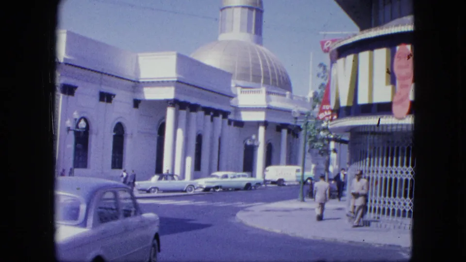 1964 Venezuela street scene with a classic car in the foreground, a white domed building, parked vehicles, pedestrians, and a striped awning.