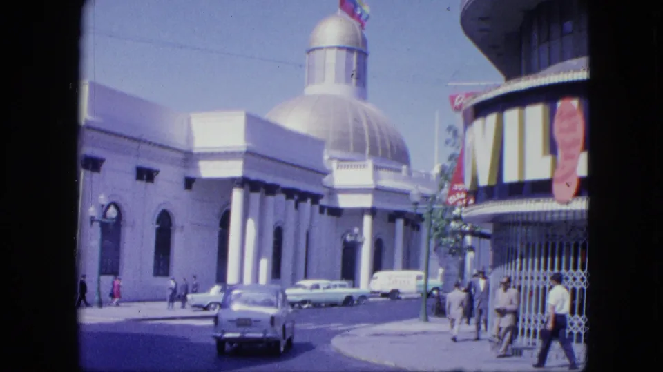 1964 Venezuela urban view with a car driving past, pedestrians on the sidewalk, parked vehicles, and a white domed building behind them.
