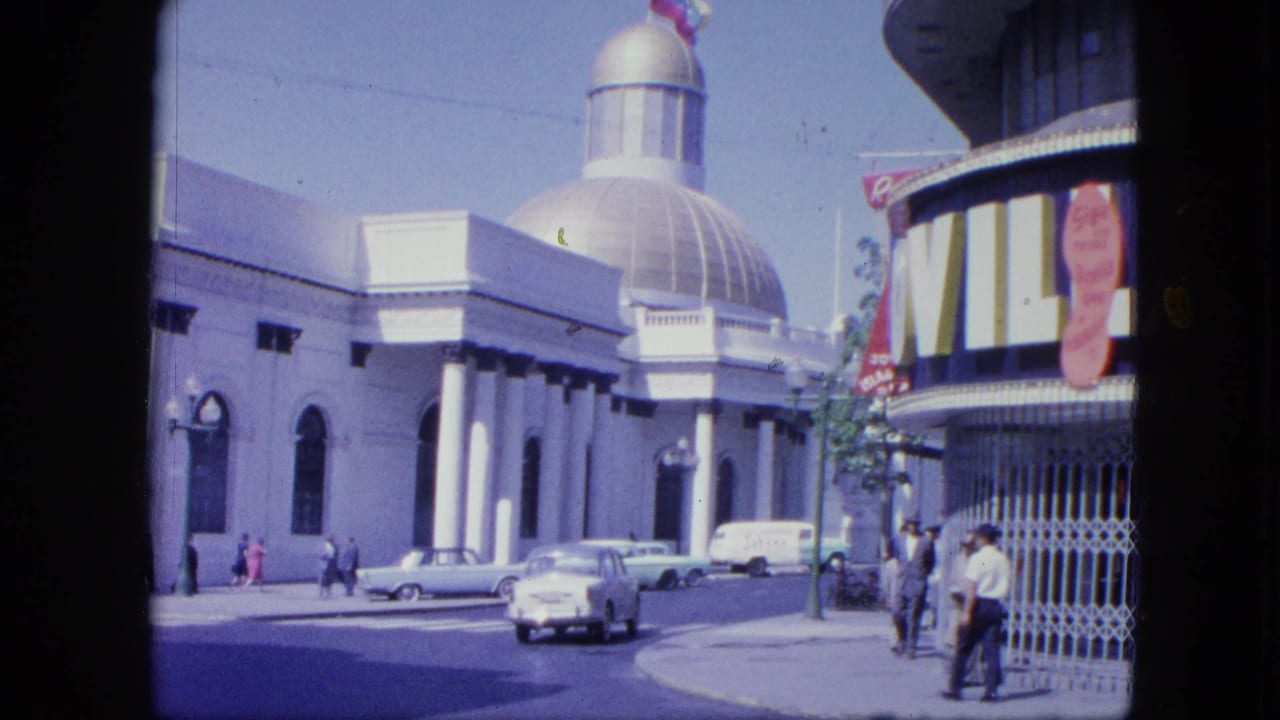 1964 Venezuela home-movie frame showing a white domed building, storefront awning, classic cars, and pedestrians on a sunny street.