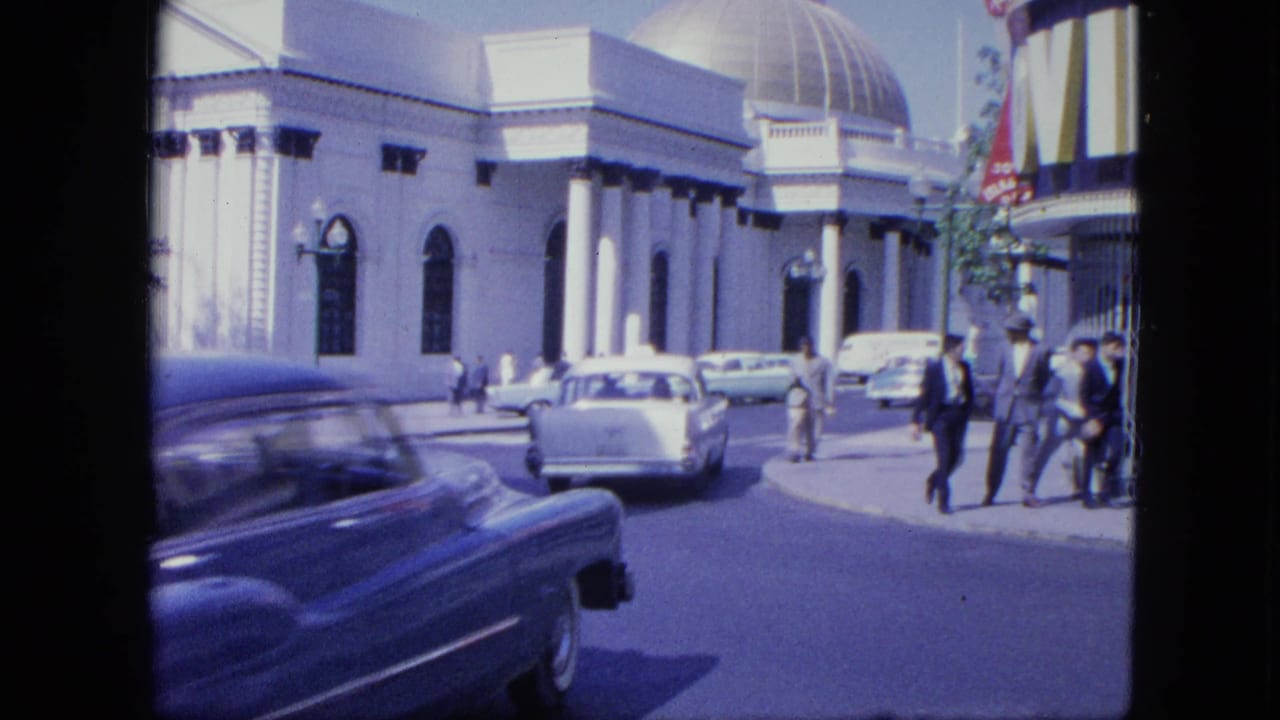 Classic car passes a white domed building as pedestrians walk nearby.