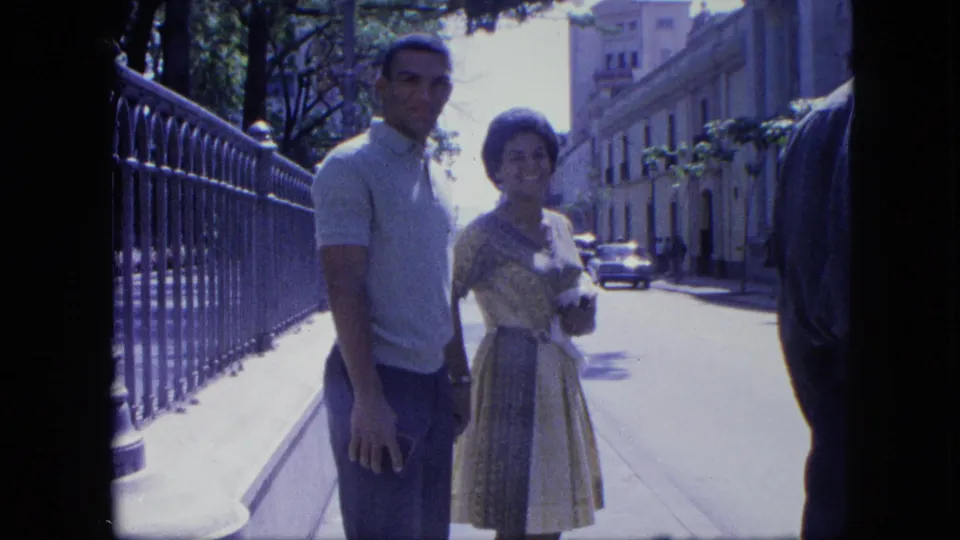 A man and woman in 1960s clothing pose on a sidewalk beside iron fencing, with cars and buildings behind them in Venezuela.