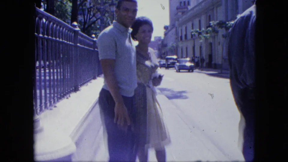 The couple remains by the iron fence on a bright Venezuelan city sidewalk, with period cars and buildings receding down the street.