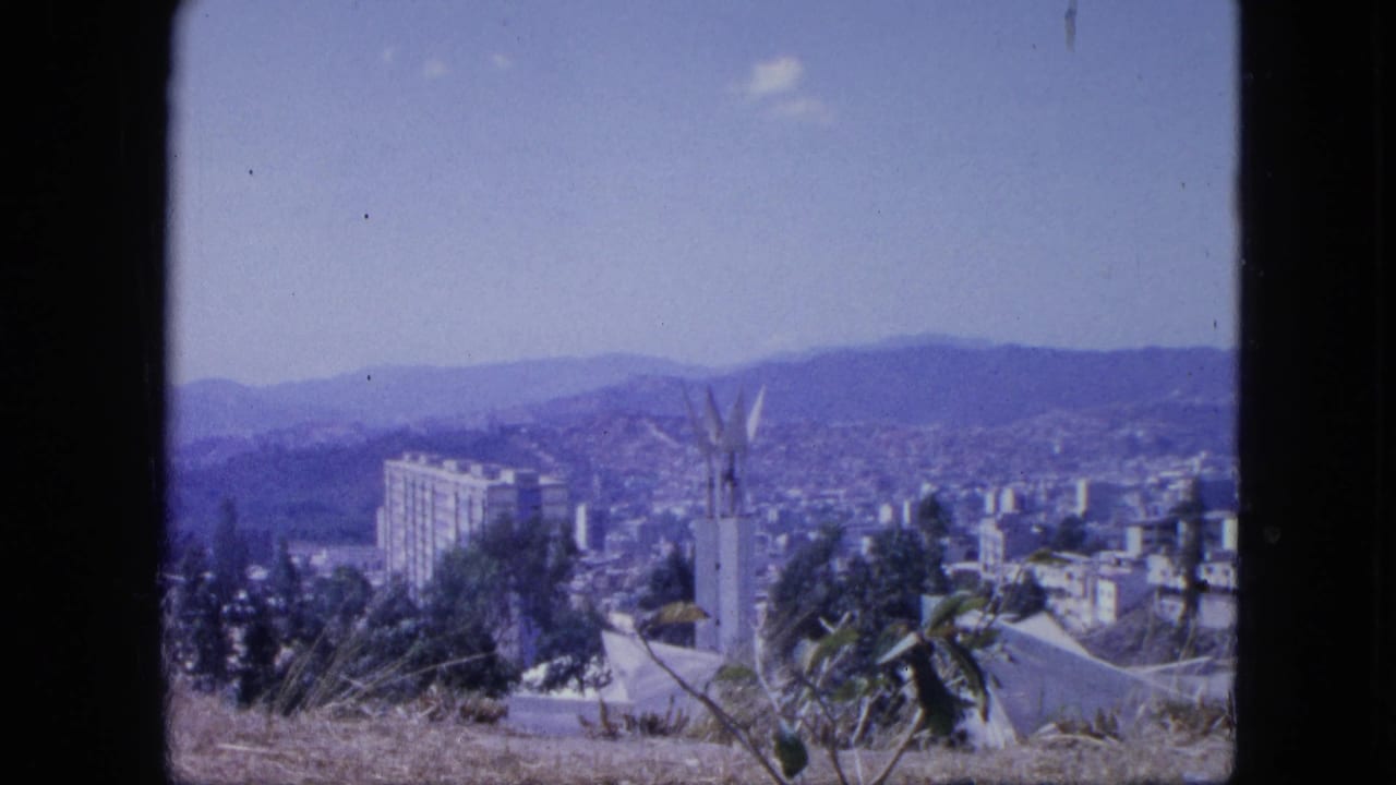1964 8mm hillside view over a Venezuelan downtown area with a tall white building, distant hills, and dry foreground plants.