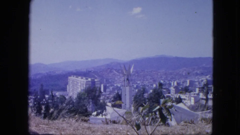 1964 8mm Venezuelan city view from a hillside, with foreground plants, a white structure, urban blocks, and distant mountain ridges.