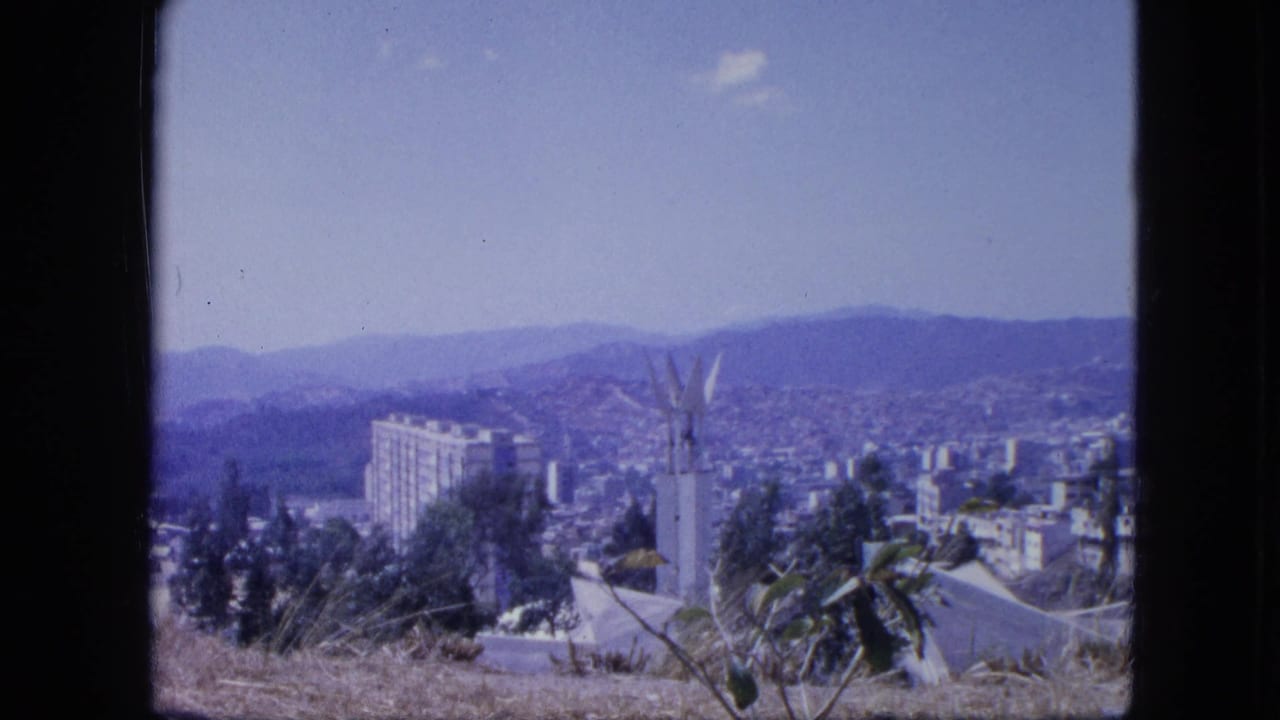 1964 Venezuela home-movie frame showing a hillside city, white monument, tall building, distant mountains, and dry foreground plants.
