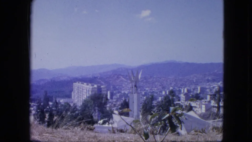 1964 Venezuela home-movie frame showing a hillside city, white monument, tall building, distant mountains, and dry foreground plants.