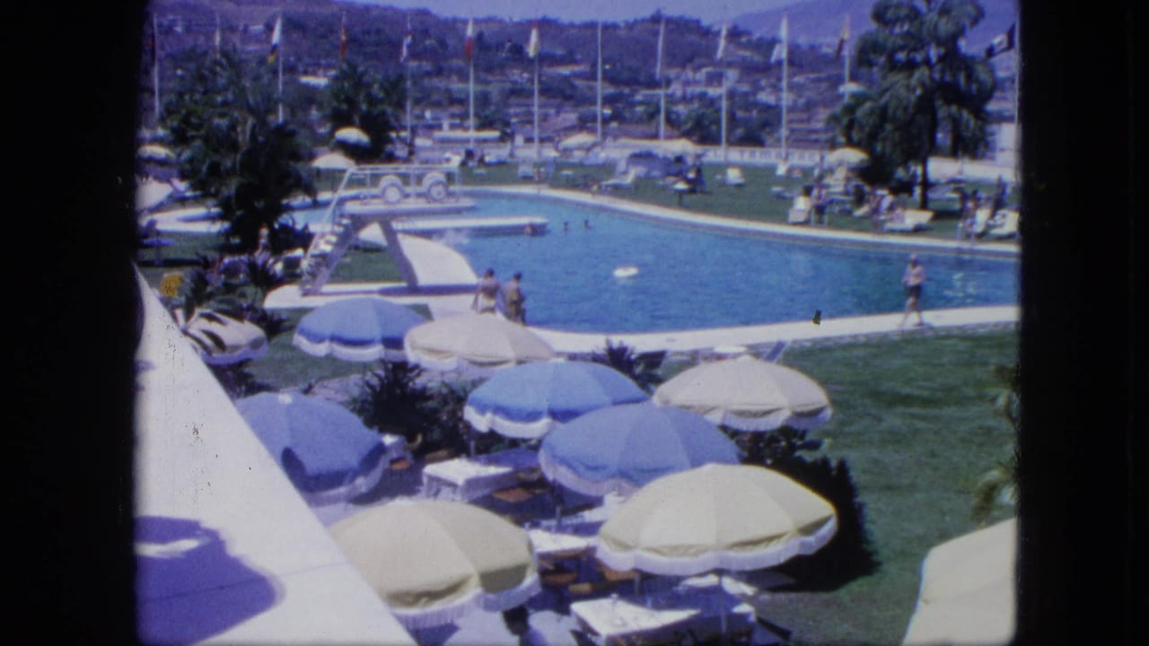 Outdoor pool with blue and white umbrellas, lounge chairs, grass, palm trees, flags, and people nearby.