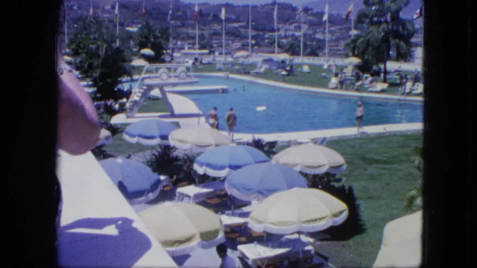 1964 Venezuela poolside frame showing swimmers, visitors, umbrellas, lounge chairs, grass, flags, palms, and a partial foreground figure.