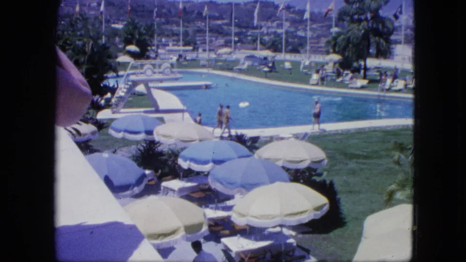 1964 Venezuela resort pool view with swimmers, blue and white umbrellas, lounge chairs, lawns, flags, palms, and hills beyond.