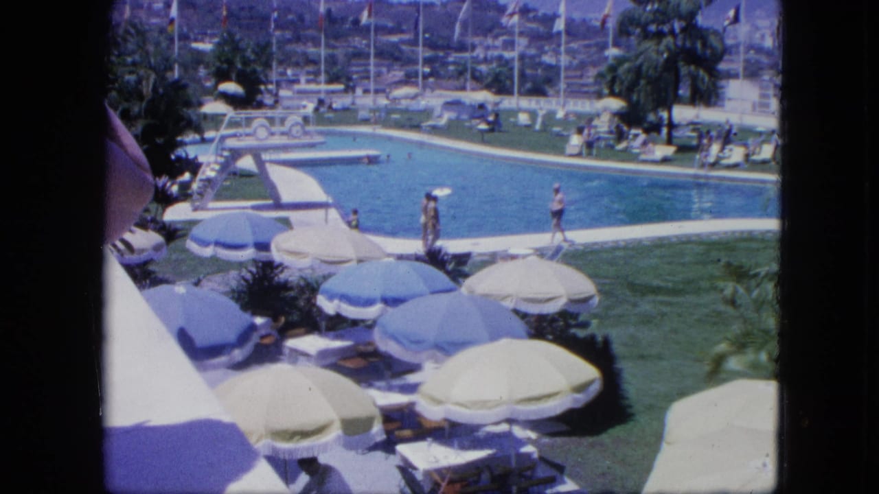 1964 8mm home-movie view of a tropical resort pool in Venezuela with guests, umbrellas, diving board, palm trees, flags, and distant hillside buildings.