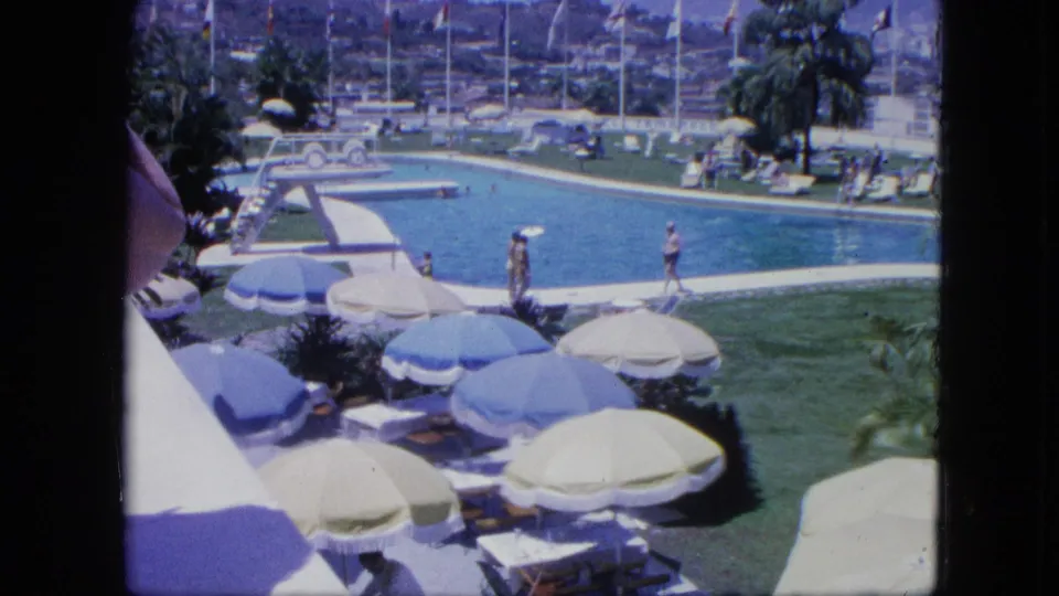 1964 8mm home-movie view of a tropical resort pool in Venezuela with guests, umbrellas, diving board, palm trees, flags, and distant hillside buildings.