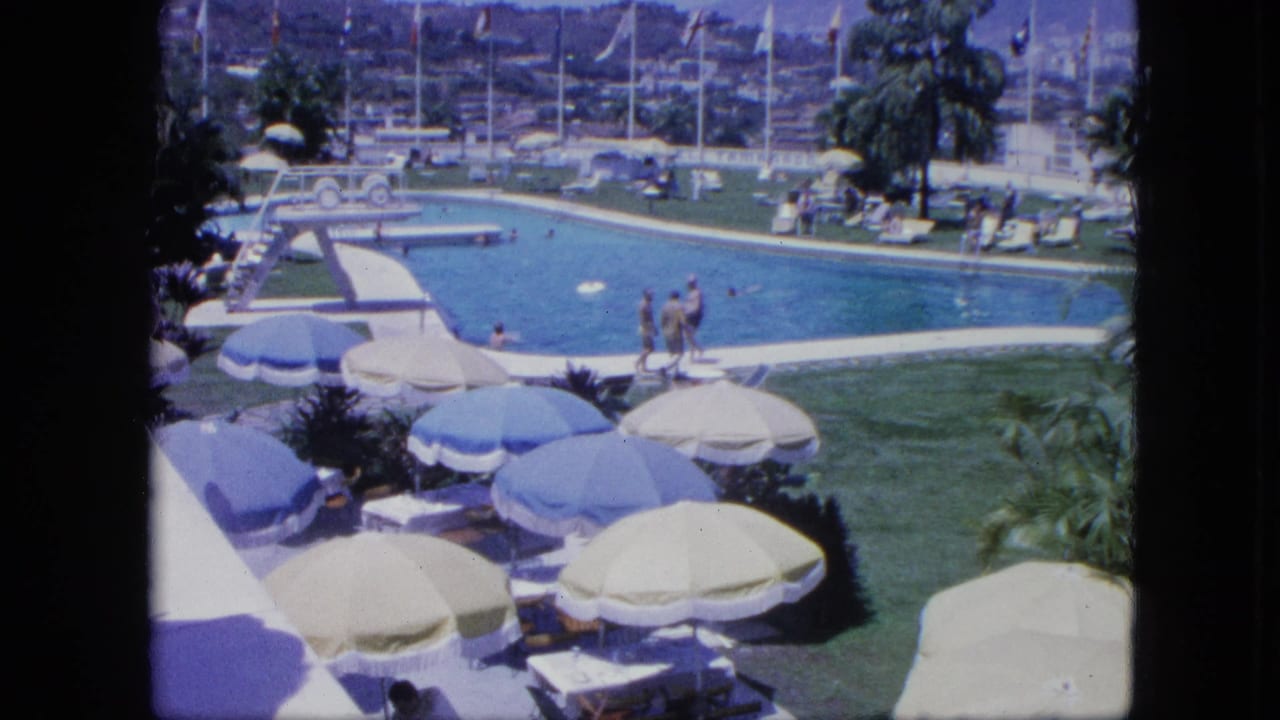 1964 8mm footage in Venezuela showing resort guests swimming and strolling near a pool bordered by umbrellas, lounge chairs, palms, and flagpoles.
