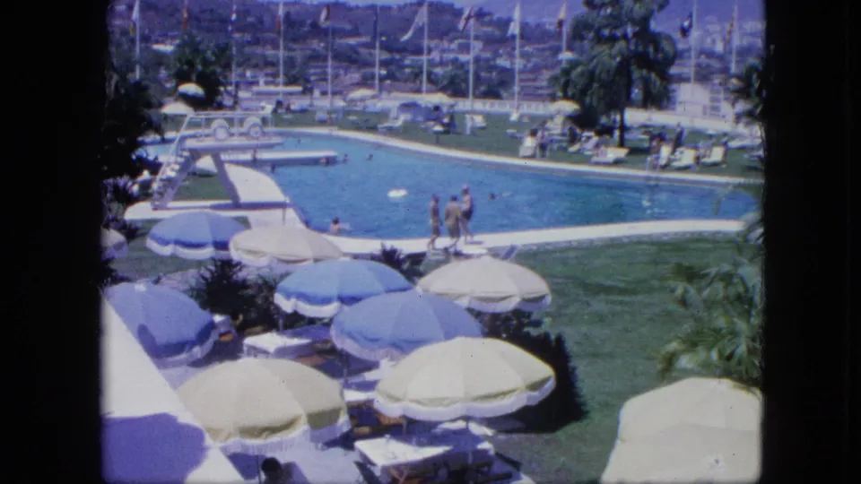 1964 8mm footage in Venezuela showing resort guests swimming and strolling near a pool bordered by umbrellas, lounge chairs, palms, and flagpoles.