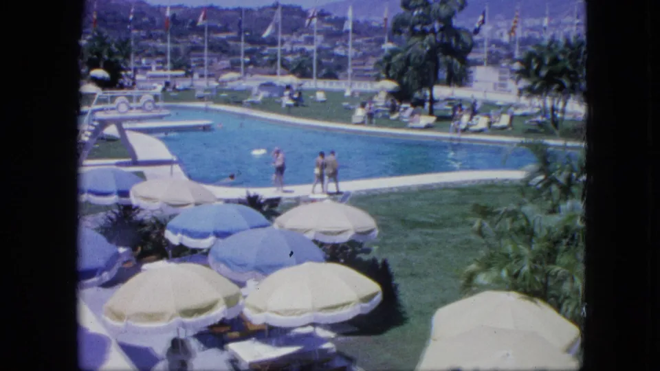 Home-movie frame from a Venezuelan resort pool area, with a diving board, guests near the water, shaded seating, palm trees, flags, and hillside development.