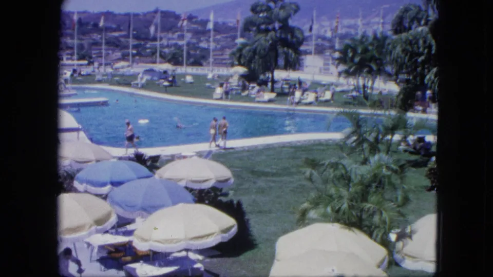 1964 Venezuela resort pool scene on 8mm film, showing swimmers and poolside guests amid umbrellas, palms, flagpoles, and distant hillside buildings.