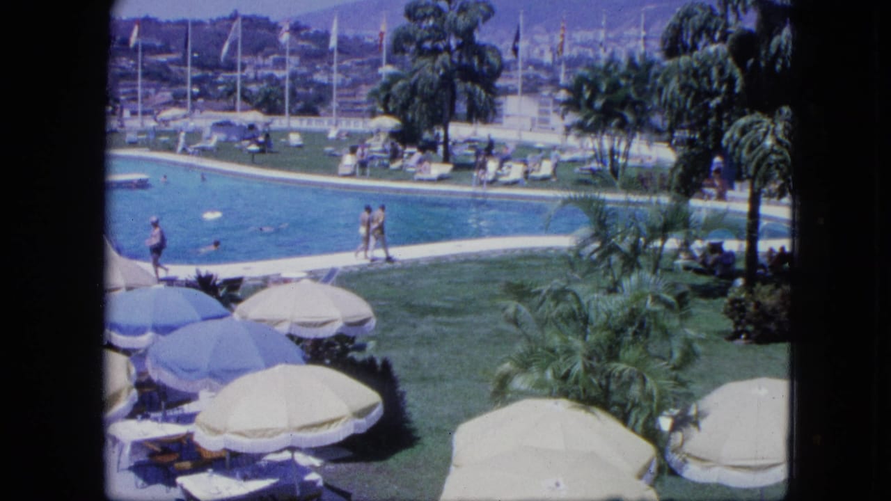 Poolside resort view with umbrellas, lawn, palm trees, guests, sailboats, and hillside buildings.