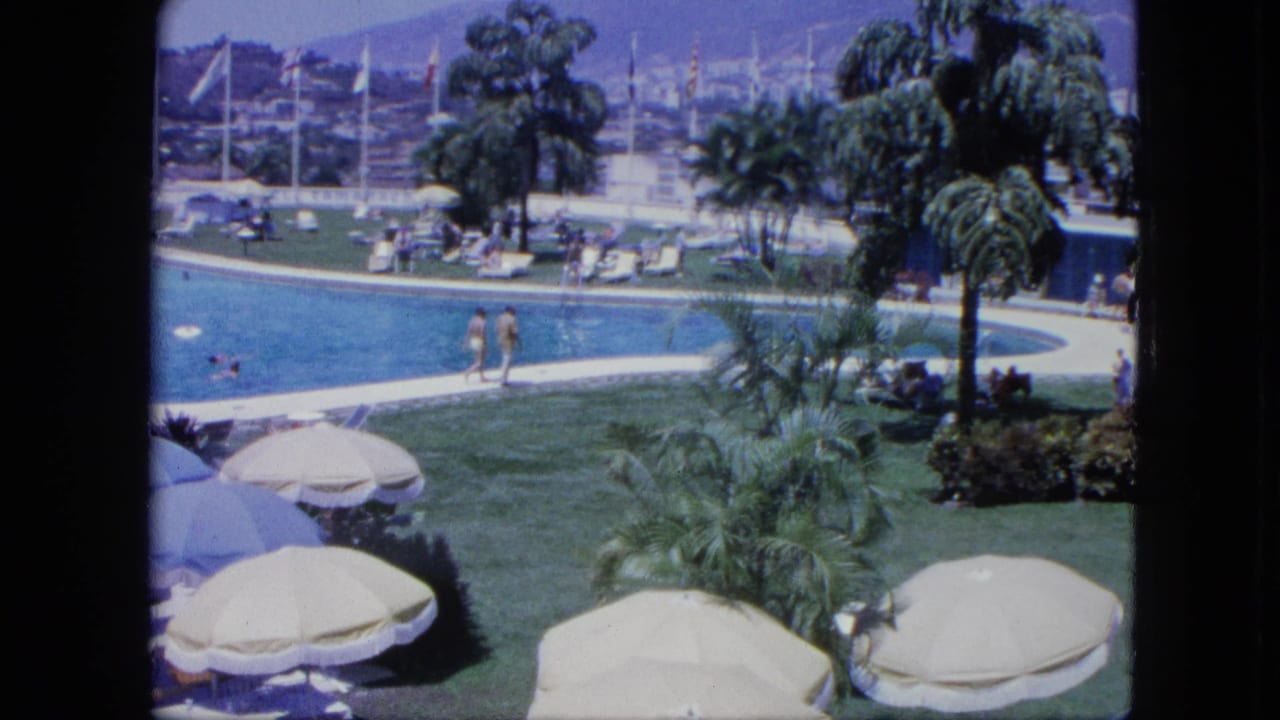 1964 Venezuela poolside resort view with two guests walking by the pool, palms and umbrellas in front, and marina masts beyond.