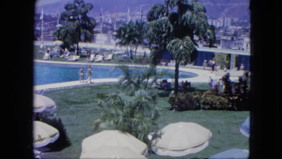 1964 Venezuela resort footage showing guests walking along the pool edge, with palms, umbrellas, trimmed lawn, and sailboats in the background.