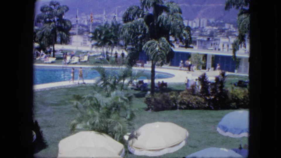 1964 Venezuela resort scene with poolside guests, tropical planting, sun umbrellas, blue water, marina masts, and hillside buildings beyond.
