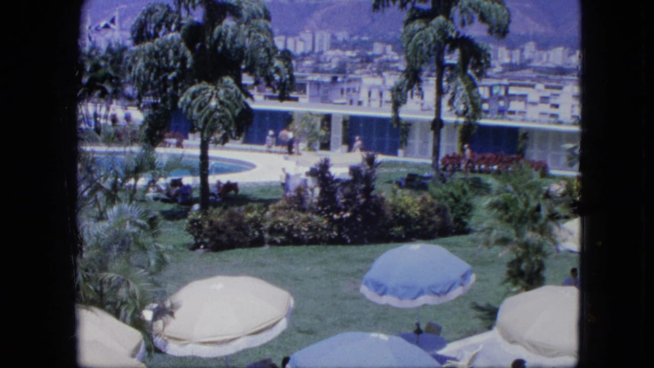 1960s home-movie frame showing resort grounds with a pool, palm trees, umbrellas, planted beds, guests, and hillside city views.