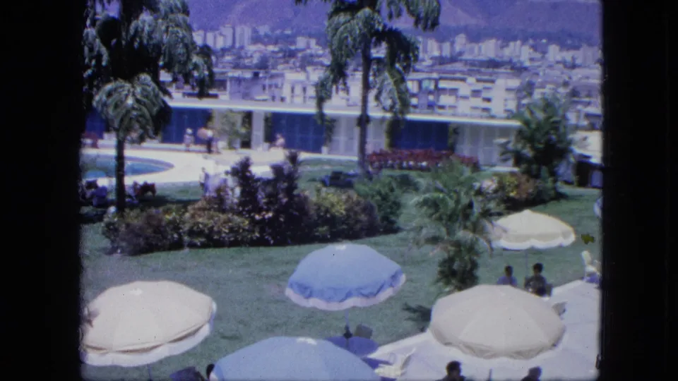 1960s 8mm resort view with umbrellas across the lawn, palm landscaping, poolside paths, seated guests, and distant urban buildings.
