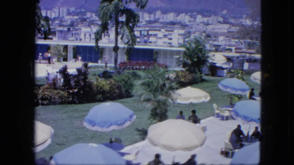1960s home-movie frame showing a wide tropical resort lawn with umbrellas, palm trees, seated guests, a pool edge, and buildings beyond.