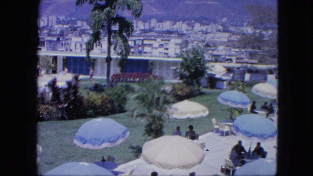 Outdoor terrace lawn with blue and white umbrellas, seated people, palms, city buildings, and mountains beyond.