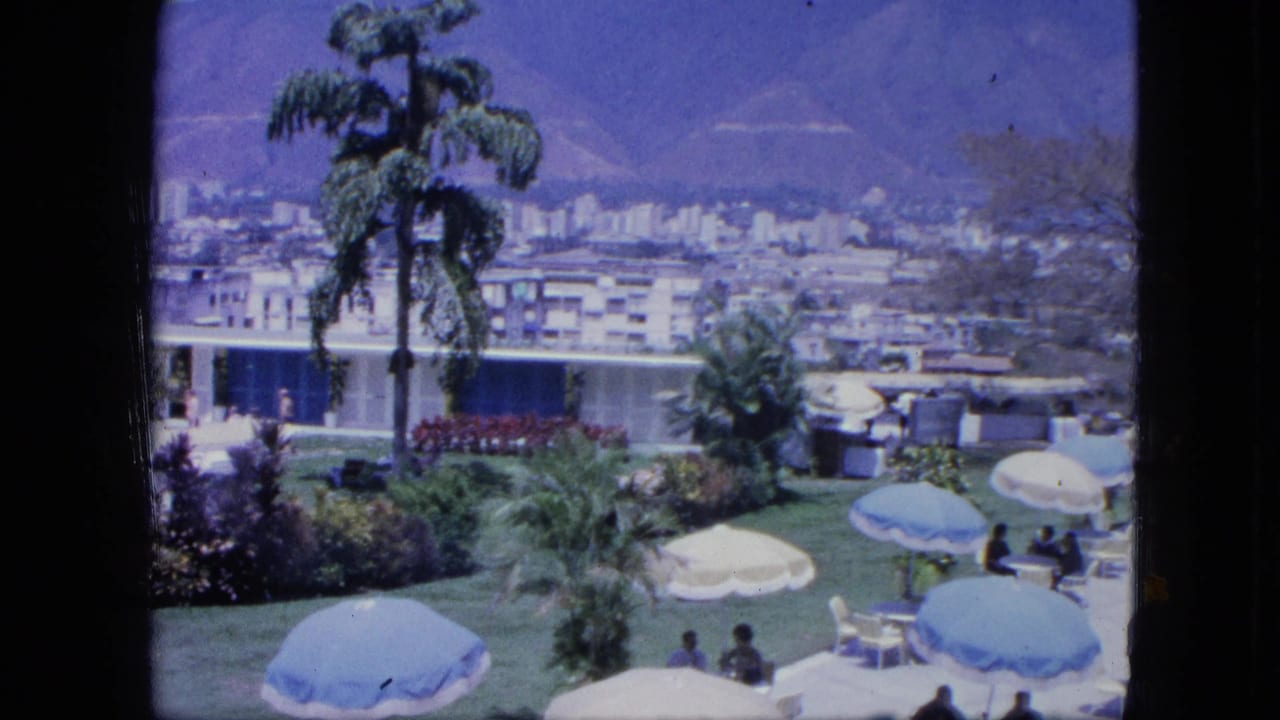Terrace lawn with umbrellas, people seated near tables, palms, planted beds, city buildings, and mountain backdrop.