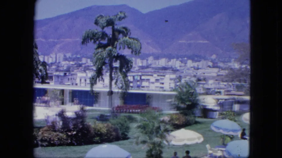 The 1964 Venezuela terrace scene shows palms and umbrellas in the foreground, seated visitors below, and a city spread against mountains.
