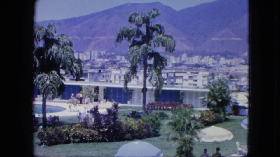 1964 Venezuela home-movie frame of a garden terrace, umbrellas, distant seated figures, a modern building, city blocks, and mountains.