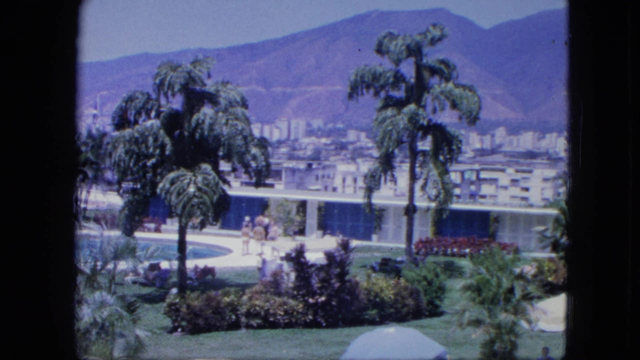 1964 8mm resort view with a swimming pool, tropical landscaping, scattered people, modern low buildings, and distant mountains.