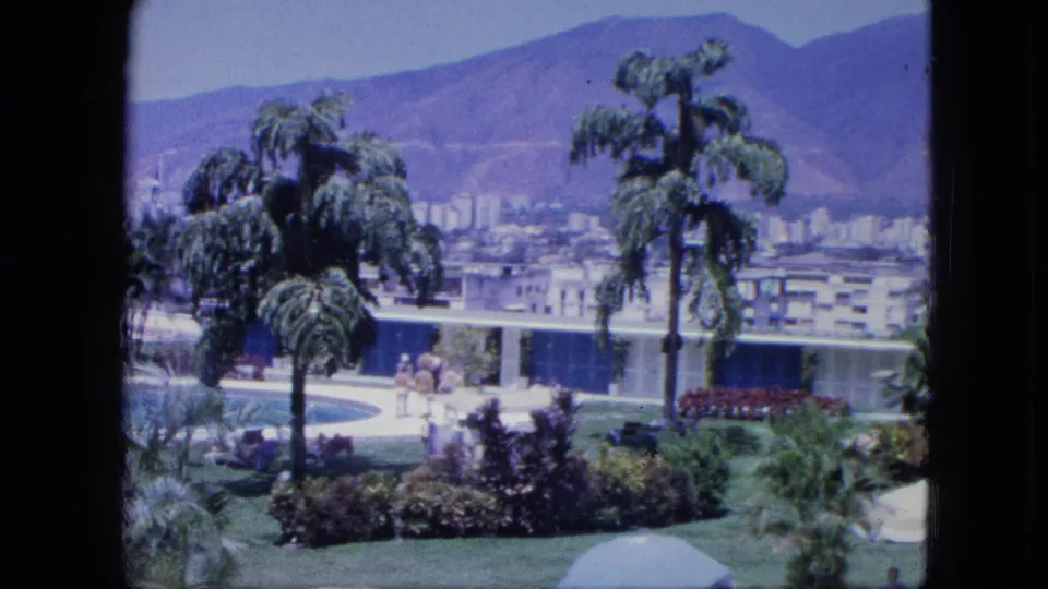 1964 8mm resort view with a swimming pool, tropical landscaping, scattered people, modern low buildings, and distant mountains.