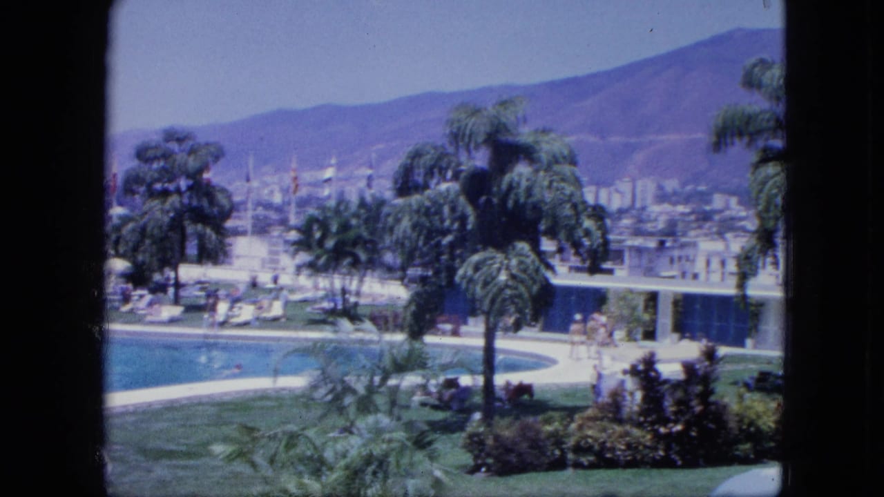 Guest-level 8mm view across a resort pool terrace with palms, lounge chairs, small figures, low buildings, and hazy mountains.