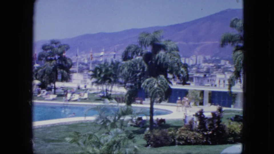 Guest-level 8mm view across a resort pool terrace with palms, lounge chairs, small figures, low buildings, and hazy mountains.