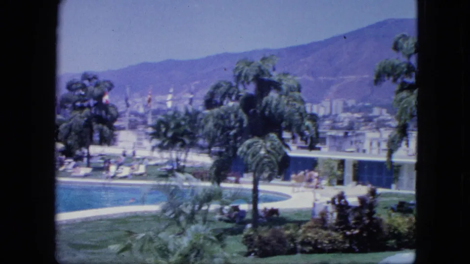 1960s 8mm frame of a tropical resort pool, green lawn, palm trees, people near the water, low buildings, and mountain views.