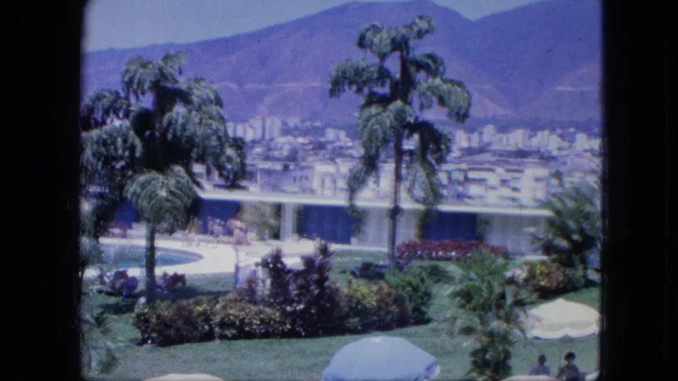 Vintage 8mm view of resort landscaping with palms, flower beds, pool edge, distant people, low modern buildings, and mountains behind.