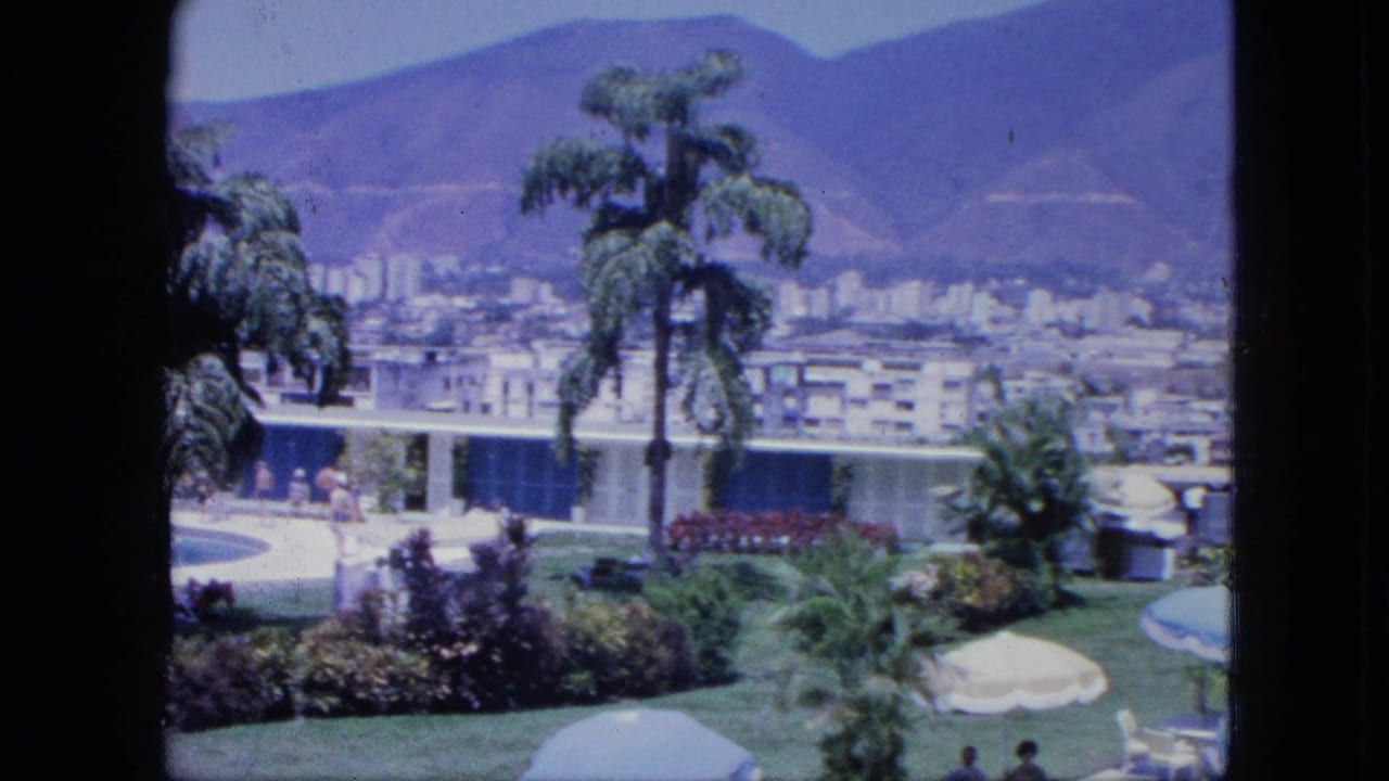 Resort terrace with palms, lawn umbrellas, pool edge, city buildings, and mountains in the background.