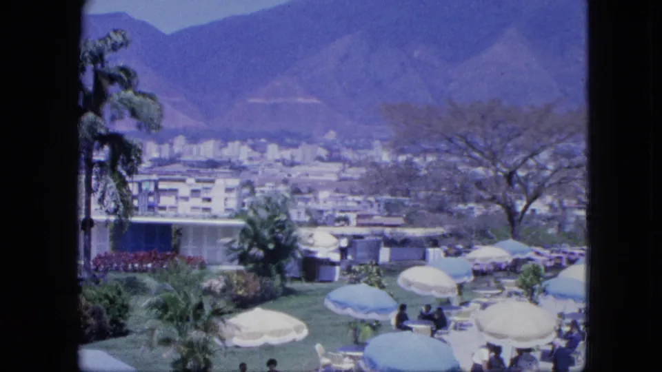 Resort lawn scene with guests under umbrellas, garden planting, parked cars, distant urban blocks, and mountains in soft 8mm color.