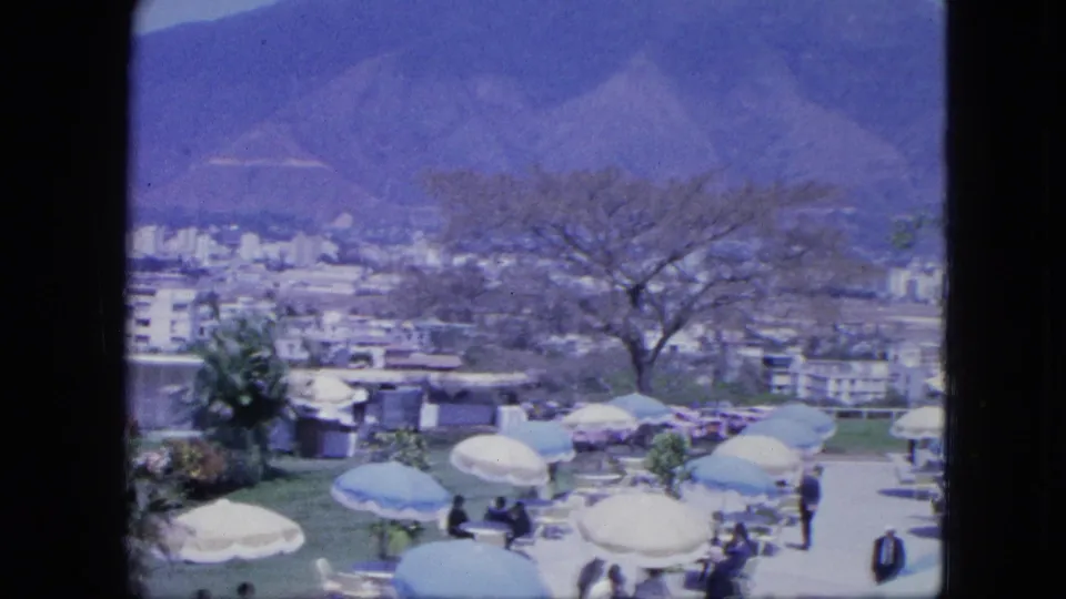 Final 8mm frame of a resort terrace with umbrellas, scattered guests, a large tree, urban buildings below, and mountains beyond.