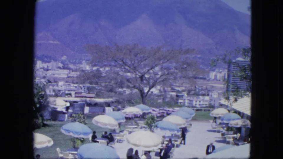 1964 home-movie frame looking down on an outdoor terrace gathering with umbrellas, seated guests, city buildings, trees, and mountains.