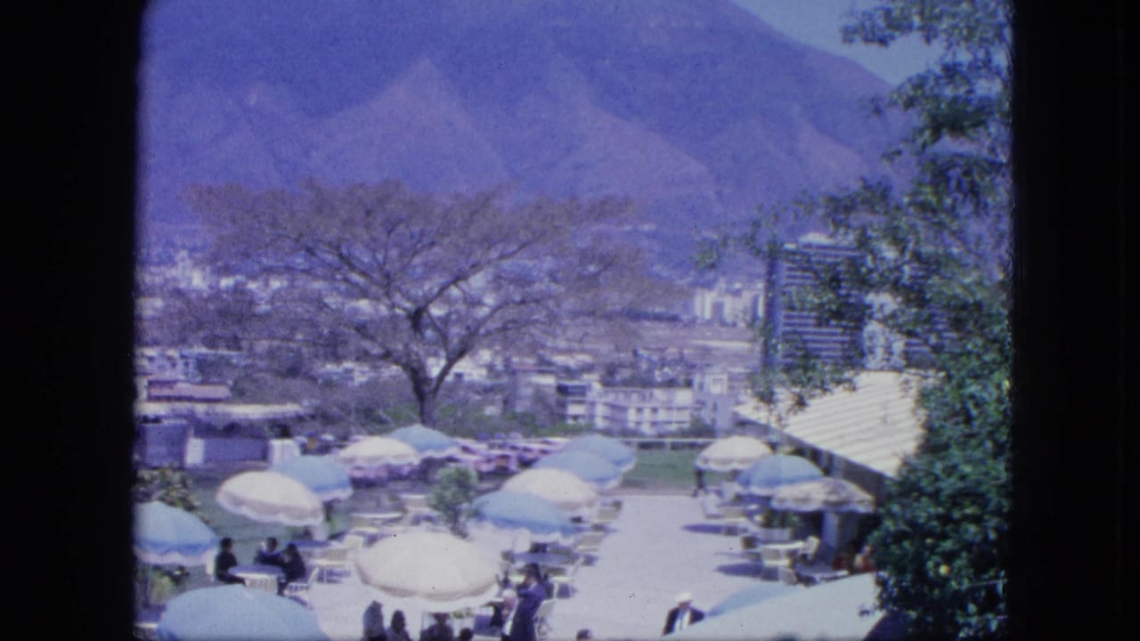 1964 home-movie view across a terrace social scene with clustered umbrellas, small groups of guests, trees, city buildings, and mountains.