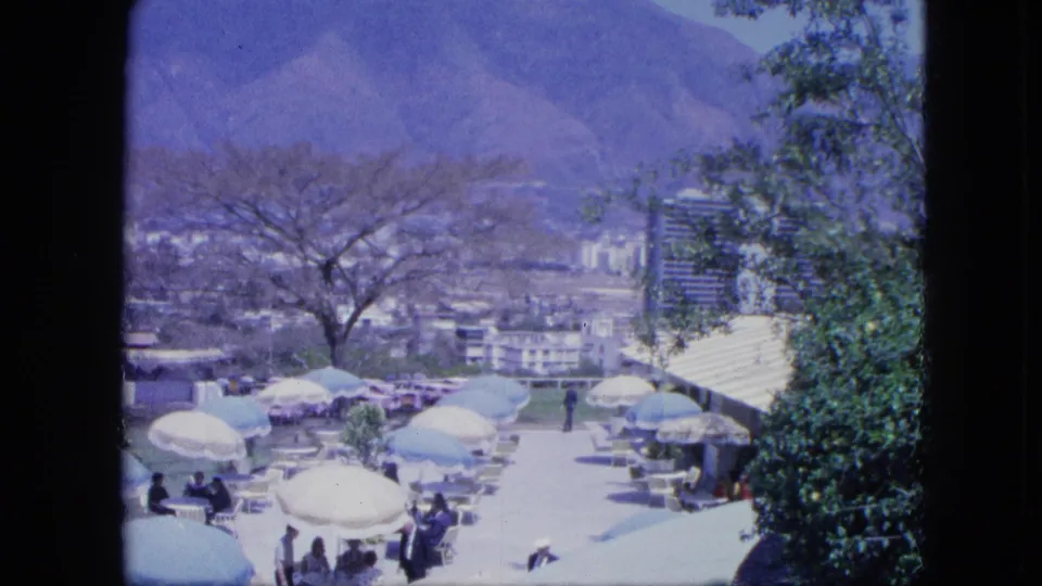 1964 home-movie frame showing a terrace party from above, with umbrellas, seated guests, greenery, city buildings, and a mountain backdrop.