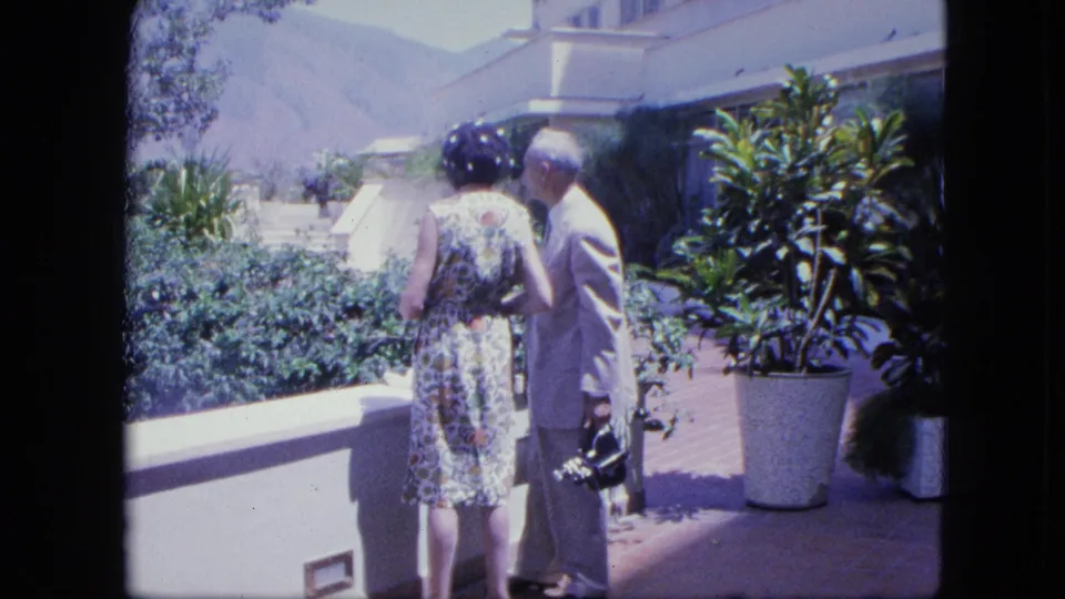 1964 home-movie frame of a woman in a patterned dress and a man in a suit standing by potted plants on a sunlit terrace.