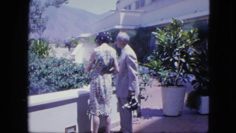 A woman in a floral dress and a man in a light suit stand on a sunlit terrace, facing away toward greenery and mountains.
