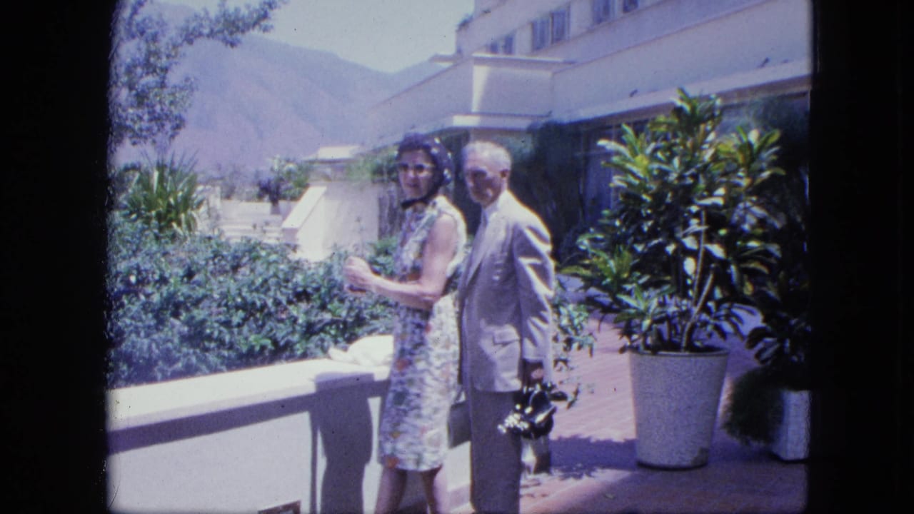 The couple has turned toward the camera on the hotel terrace, with the man holding a camera and mountains in the background.