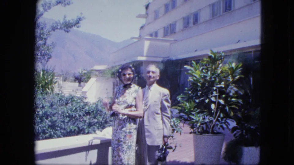 A smiling couple stands together on the terrace, framed by potted plants, a hotel facade, and a hazy mountain slope.